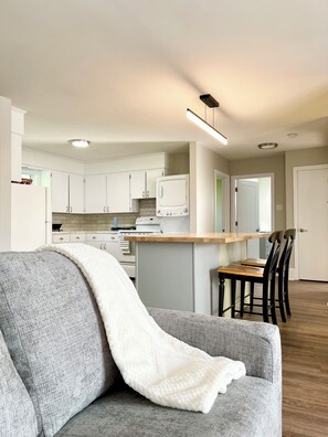 A modern kitchen with white cabinets, a built-in oven, and a microwave. There is a dining area with a wooden table and black chairs.