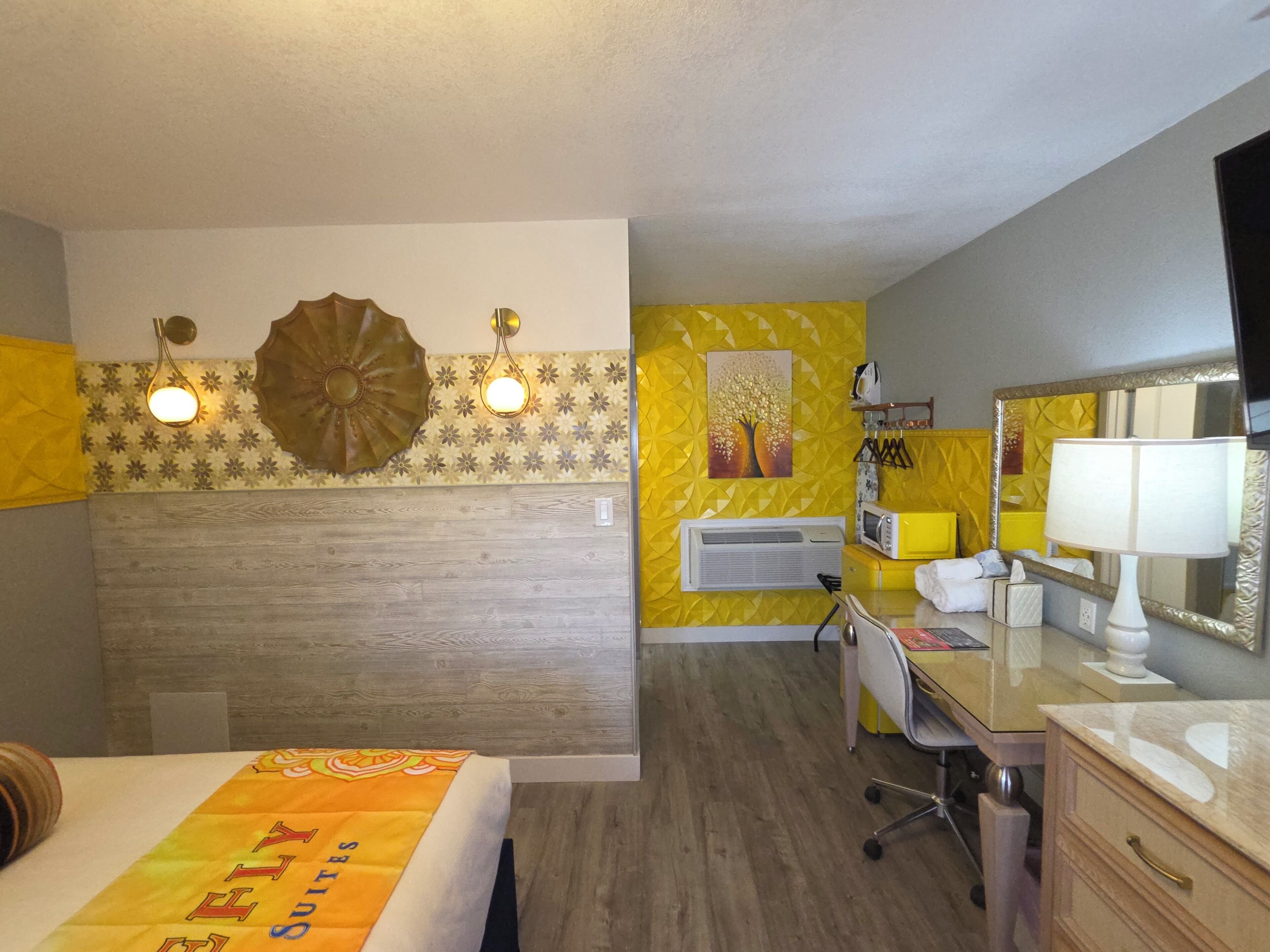 A hotel room with a bed, a desk, a chair, a mirror, a lamp, and a wall-mounted air conditioner.