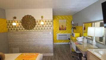A hotel room with a bed, a desk, a chair, a mirror, a lamp, and a wall-mounted air conditioner.