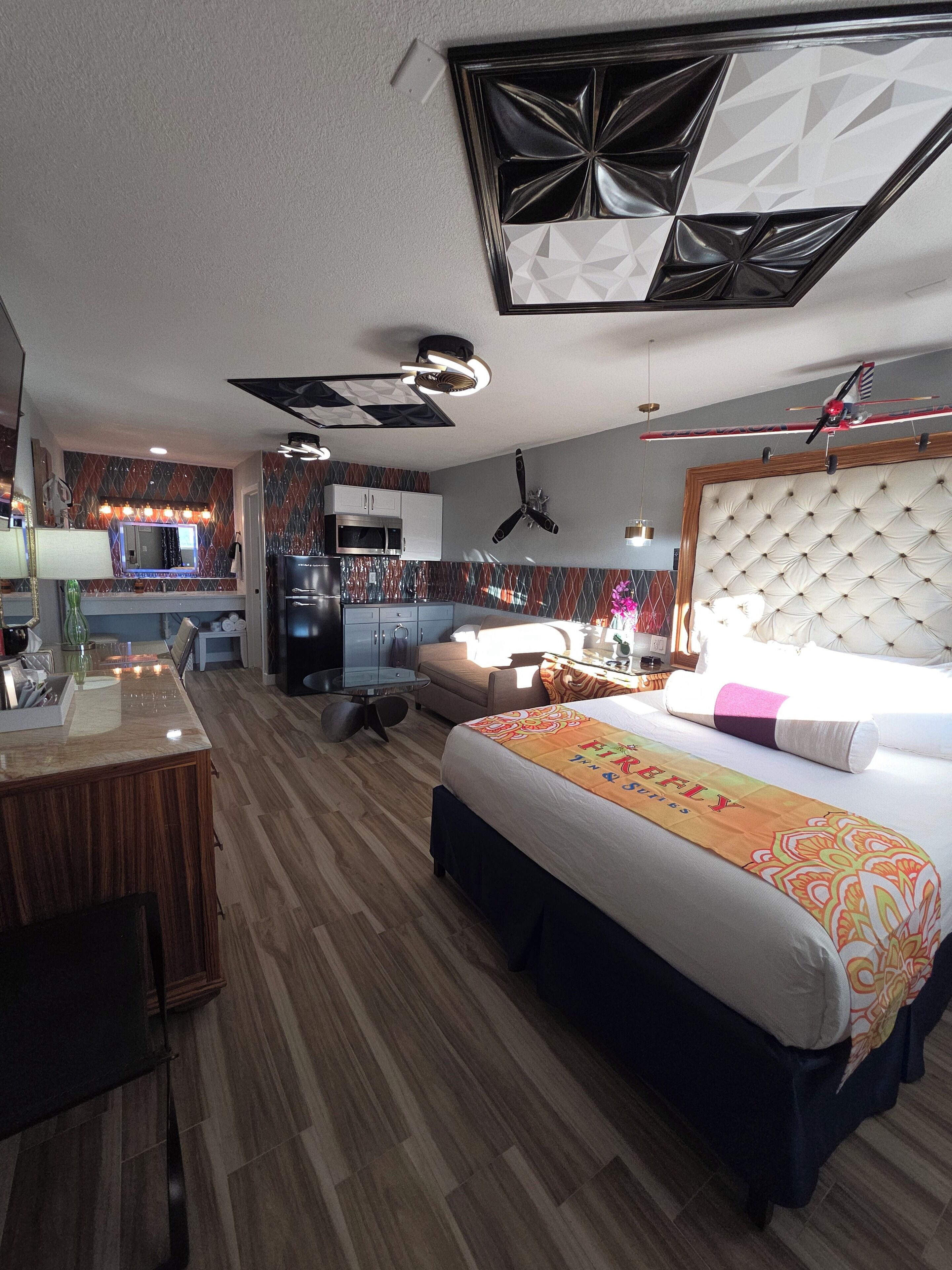 A hotel room with a bed, a desk, a kitchenette, and a ceiling fan.