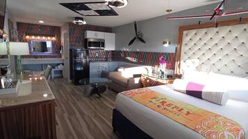 A hotel room with a bed, a desk, a kitchenette, and a ceiling fan.