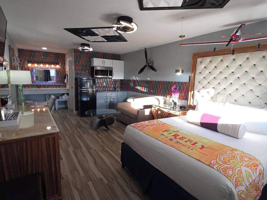 A hotel room with a bed, a desk, a kitchenette, and a ceiling fan.