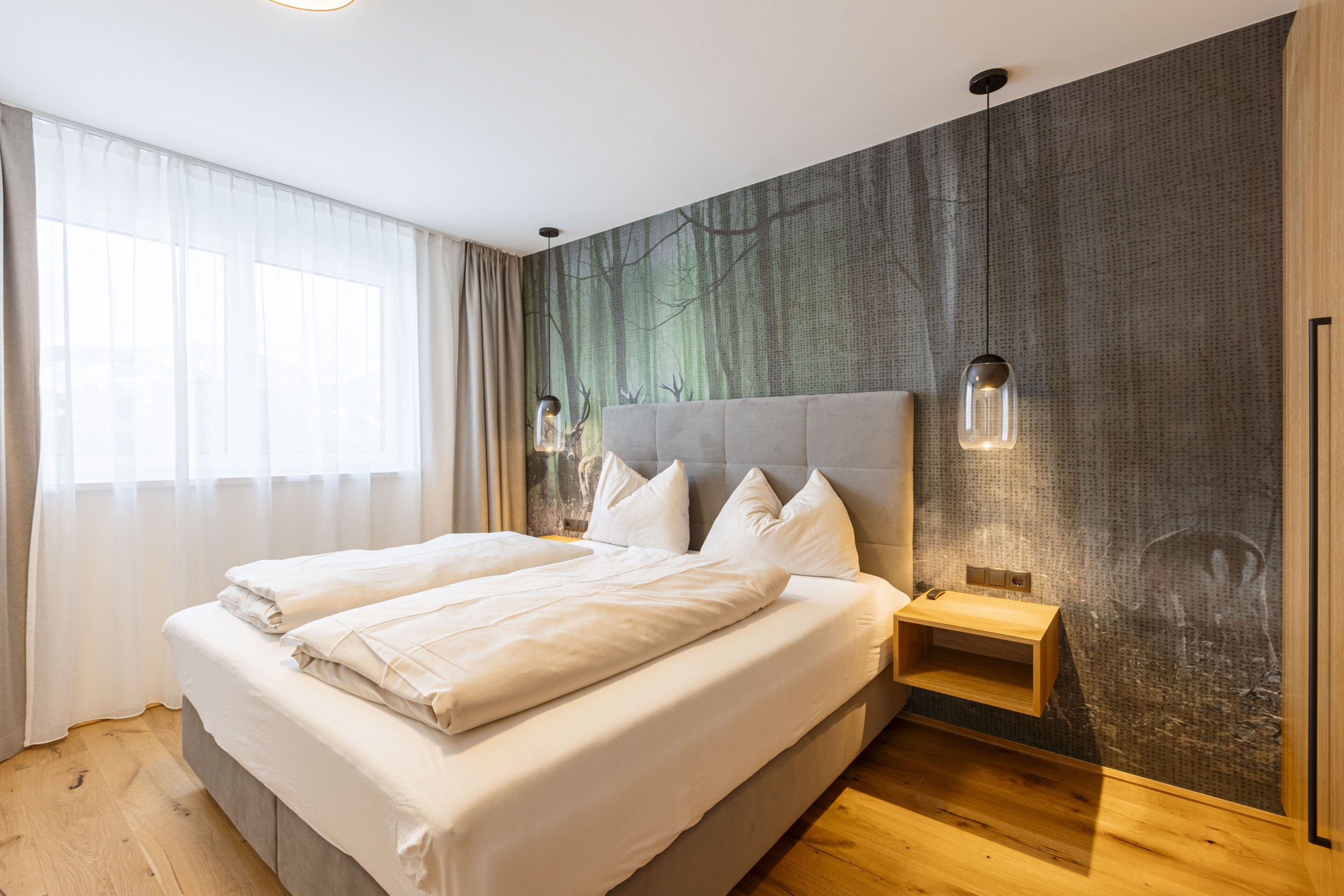 A modern bedroom with a large bed, a nightstand, a wall-mounted lamp, and a window with sheer curtains.
