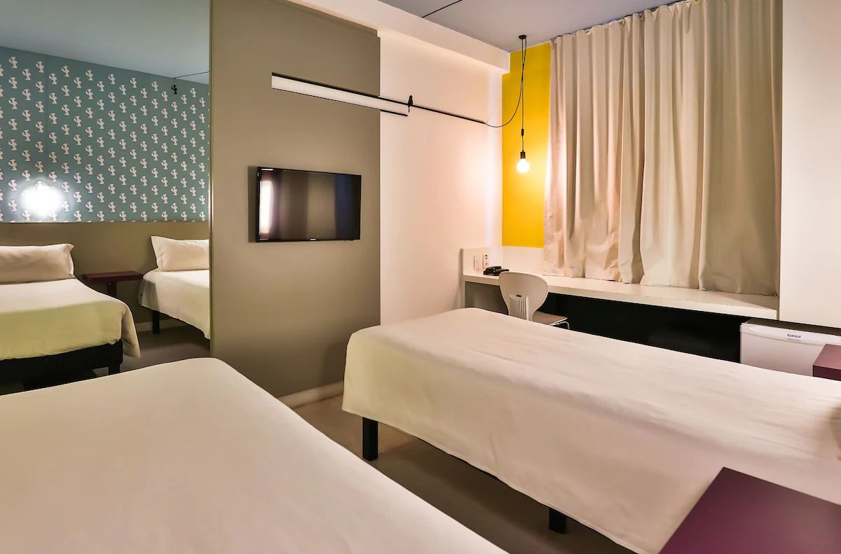 Quarto de hotel com duas camas, televisão, escrivaninha e janela com cortinas.