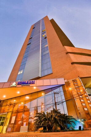 Ein hohes Gebäude mit modernem Design, das eine auffällige Eingangszone mit Glasüren und einem Schild mit der Aufschrift "NOBLE INN PAMPULHA" zeigt.