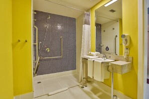 Ein Badezimmer mit gelber Wand, einer Dusche mit Glastür, einem Waschbecken mit Spiegel und einem an der Wand befestigten Föhn.