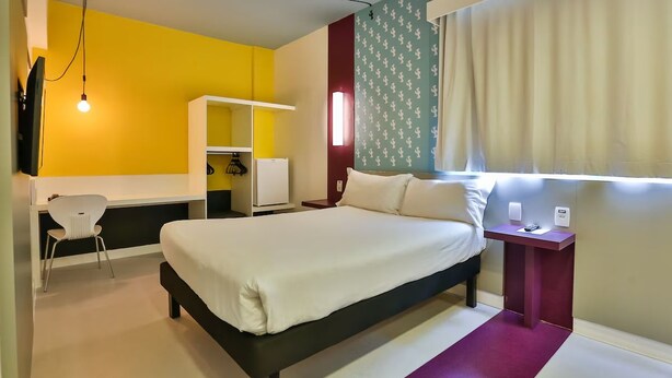 Habitación de hotel con cama, escritorio, silla, armario y ventana con cortinas.