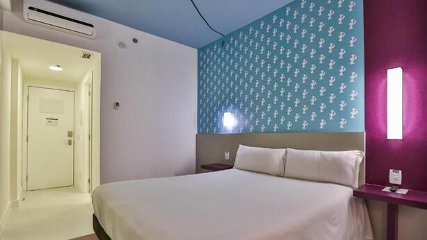 Una camera d'albergo con un letto, un comodino, una lampada a parete e una porta che dà su un'altra stanza.