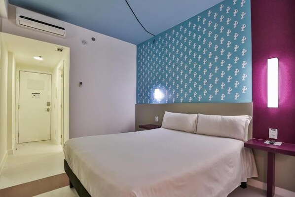 Una habitación de hotel con una cama, una mesita de noche, una lámpara fijada a la pared y una puerta que da a otra habitación.