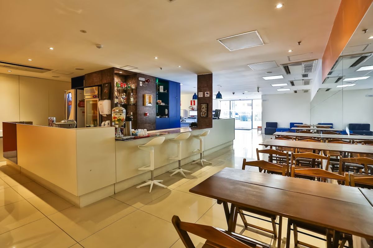Uma área de refeições moderna com um bar central, banquetas brancas e mesas de madeira.