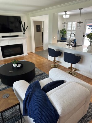 Ein modernes Wohnzimmer mit Kamin, einem runden schwarzen Couchtisch, einer blauen Sitzsofa und einer Küchenzeile mit Bar und Pendelleuchten.