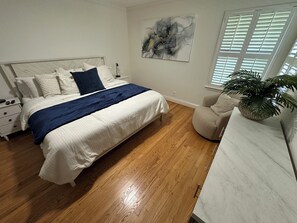 Ein Schlafzimmer mit einem Bett, Nachttischen, einem Sessel, einer Zimmerpflanze und einem Fenster mit Jalousien.