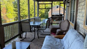 A covered porch with a wooden table, chairs, a sofa, and a view of a house and trees. - Waterfront lake house retreat located in Baileyville Maine! (Baileyville)