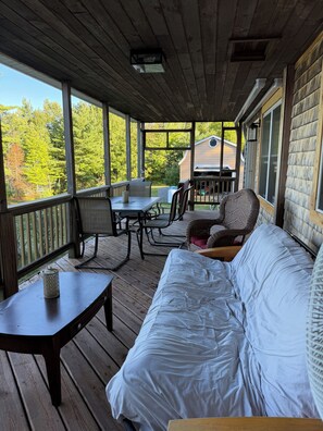 A covered porch with a wooden table, chairs, a sofa, and a view of a house and trees. - Waterfront lake house retreat located in Baileyville Maine! (Baileyville)