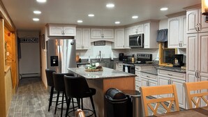 A modern kitchen with white cabinetry, a central island with bar stools, and a dining table with chairs. - Waterfront lake house retreat located in Baileyville Maine! (Baileyville)