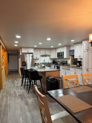 A modern kitchen with white cabinetry, a central island with bar stools, and a dining table with chairs. - Waterfront lake house retreat located in Baileyville Maine! (Baileyville)