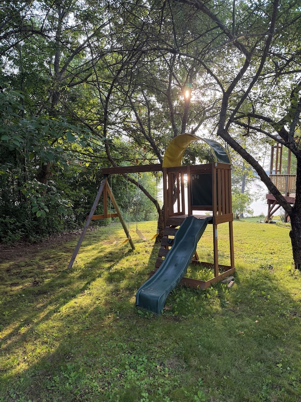 A playground with a slide and swings, surrounded by trees and grass.