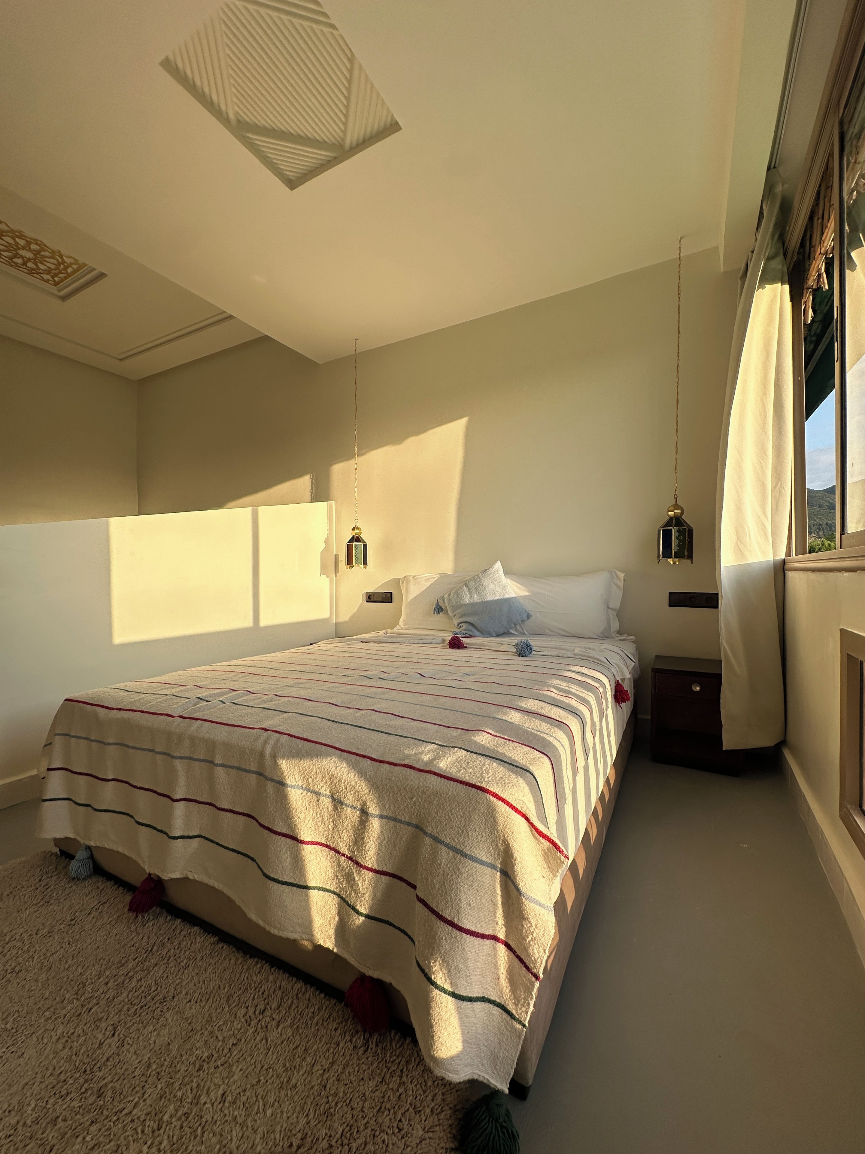 A bedroom with a bed, a nightstand, a hanging lantern, and a window with curtains.