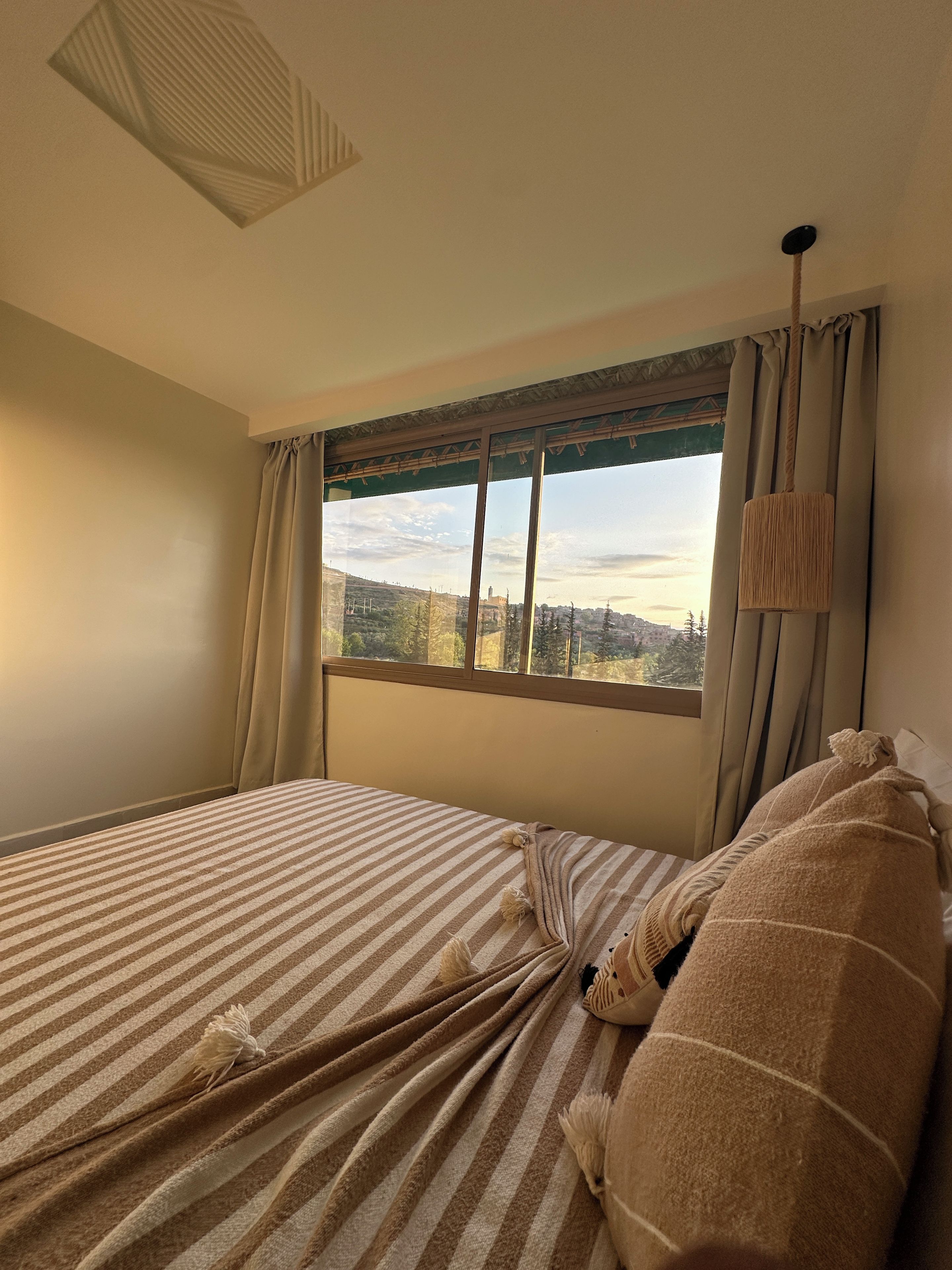 A hotel room with a large window, a bed with striped bedding, and a view of a cityscape.