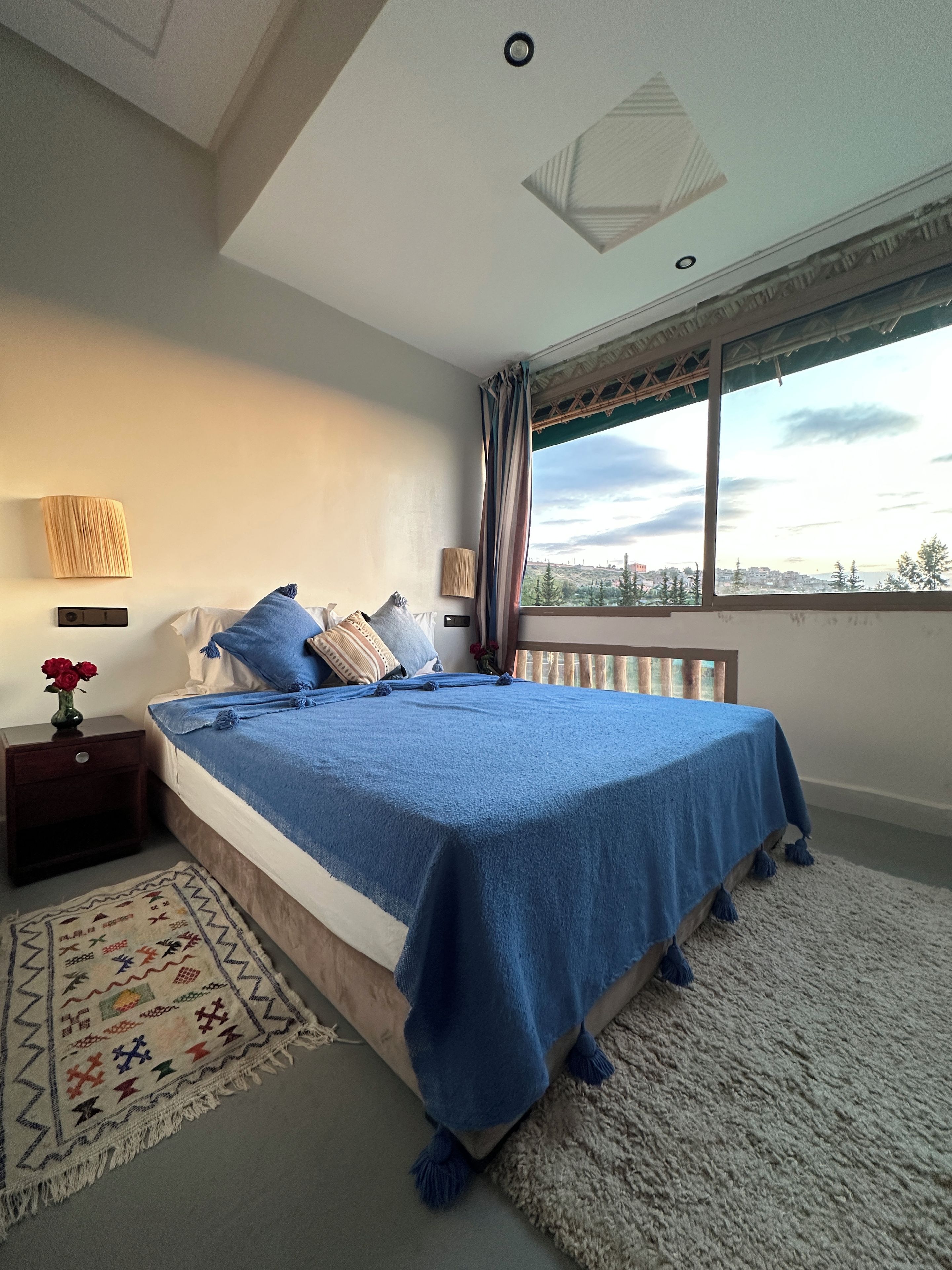 A bedroom with a bed, nightstand, lamp, and a view of the city through a window.