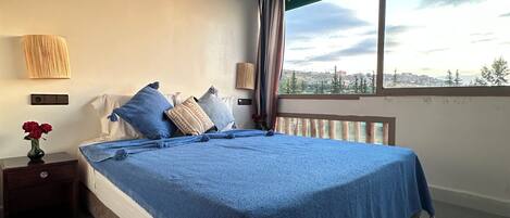 A bedroom with a bed, nightstand, lamp, and a view of the city through a window.