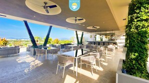 An outdoor dining area with multiple tables and chairs, ceiling fans, and a view of the landscape. - Brand New Condo Brisa Nayar 204 - Rooftop Pool & Ocean Views (Las Jarretaderas)