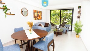 A modern living room with a round wooden table, blue upholstered chairs, a sofa, and a large window with a view of trees. - Brand New Condo Brisa Nayar 204 - Rooftop Pool & Ocean Views (Las Jarretaderas)