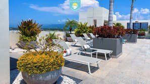 A rooftop terrace with lounge chairs, potted plants, and a view of the ocean. - Brand New Condo Brisa Nayar 204 - Rooftop Pool & Ocean Views (Las Jarretaderas)