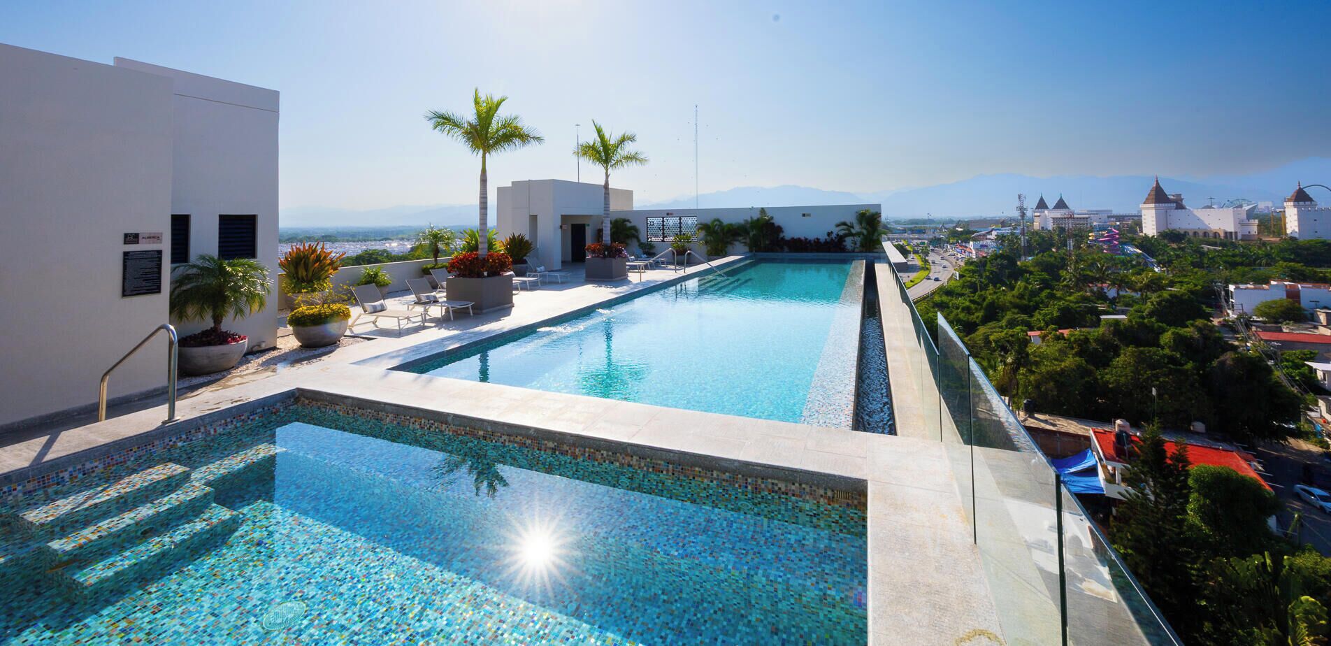 Piscina com borda azulejada, rodeada por espreguiçadeiras e plantas em vasos, com vista para a paisagem urbana e montanhas ao longe.