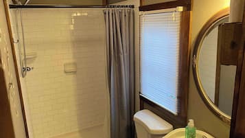A bathroom with a shower, toilet, sink, and a window with blinds.