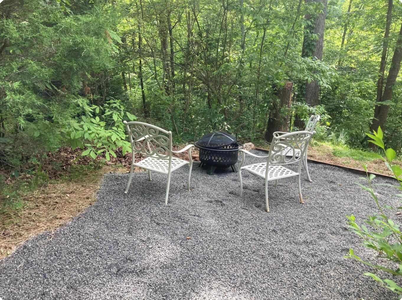 A gravel patio with two white metal chairs and a black fire pit in a forested area.