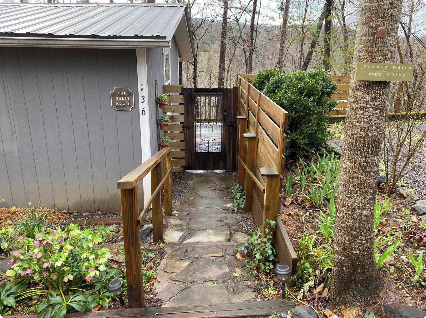 A wooden pathway leading to a guest house with a metal roof, surrounded by greenery and a wooden fence.