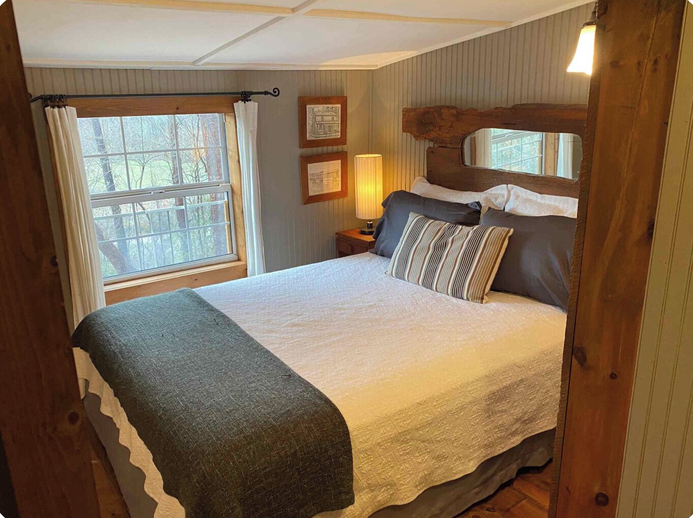 Un dormitorio con cama, ventana con cortinas, mesita de noche con lámpara y pared con cuadros enmarcados.