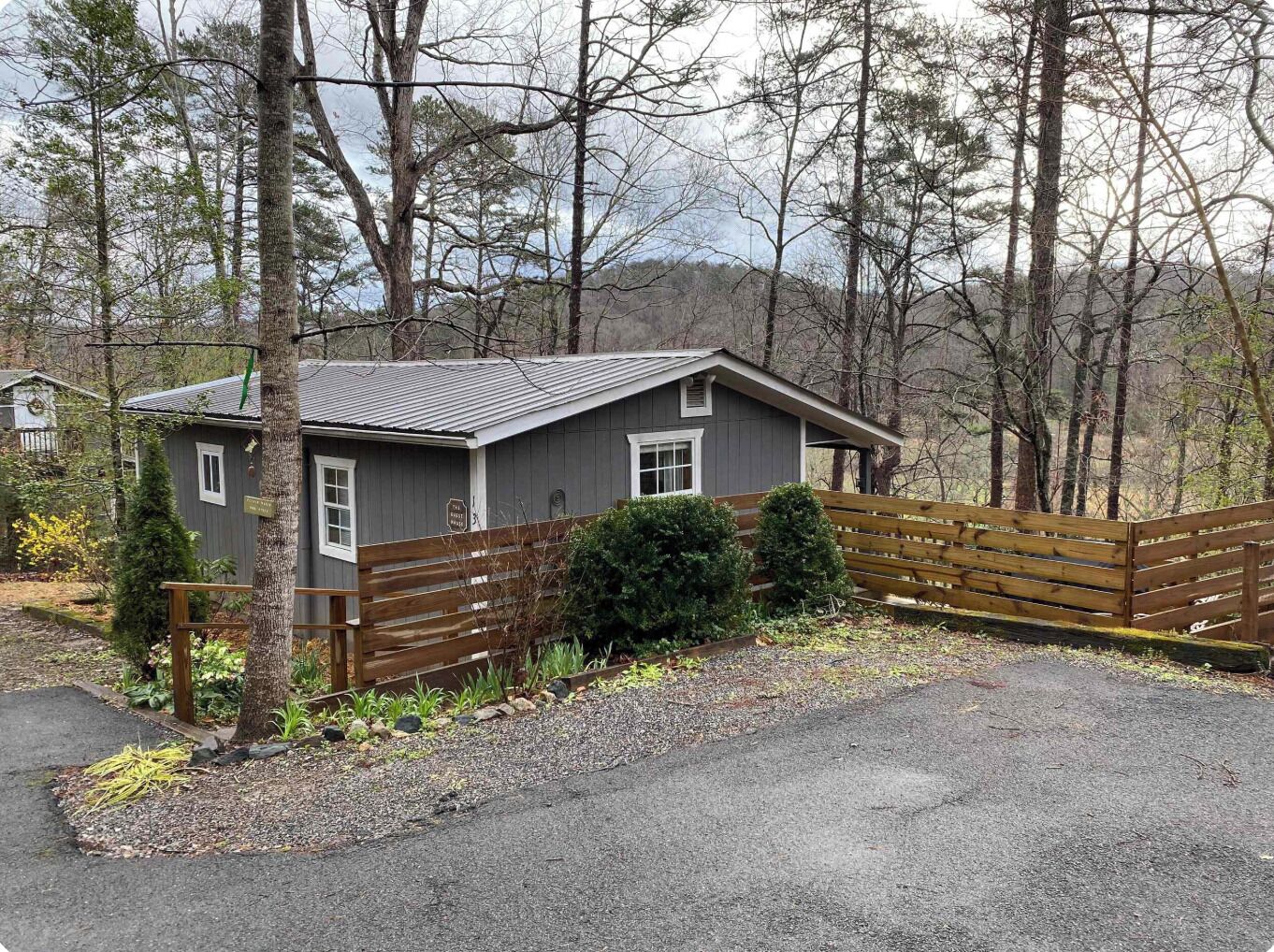 A small cabin with a wooden deck, surrounded by trees and a gravel driveway.