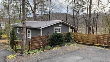 A small cabin with a wooden deck, surrounded by trees and a gravel driveway.