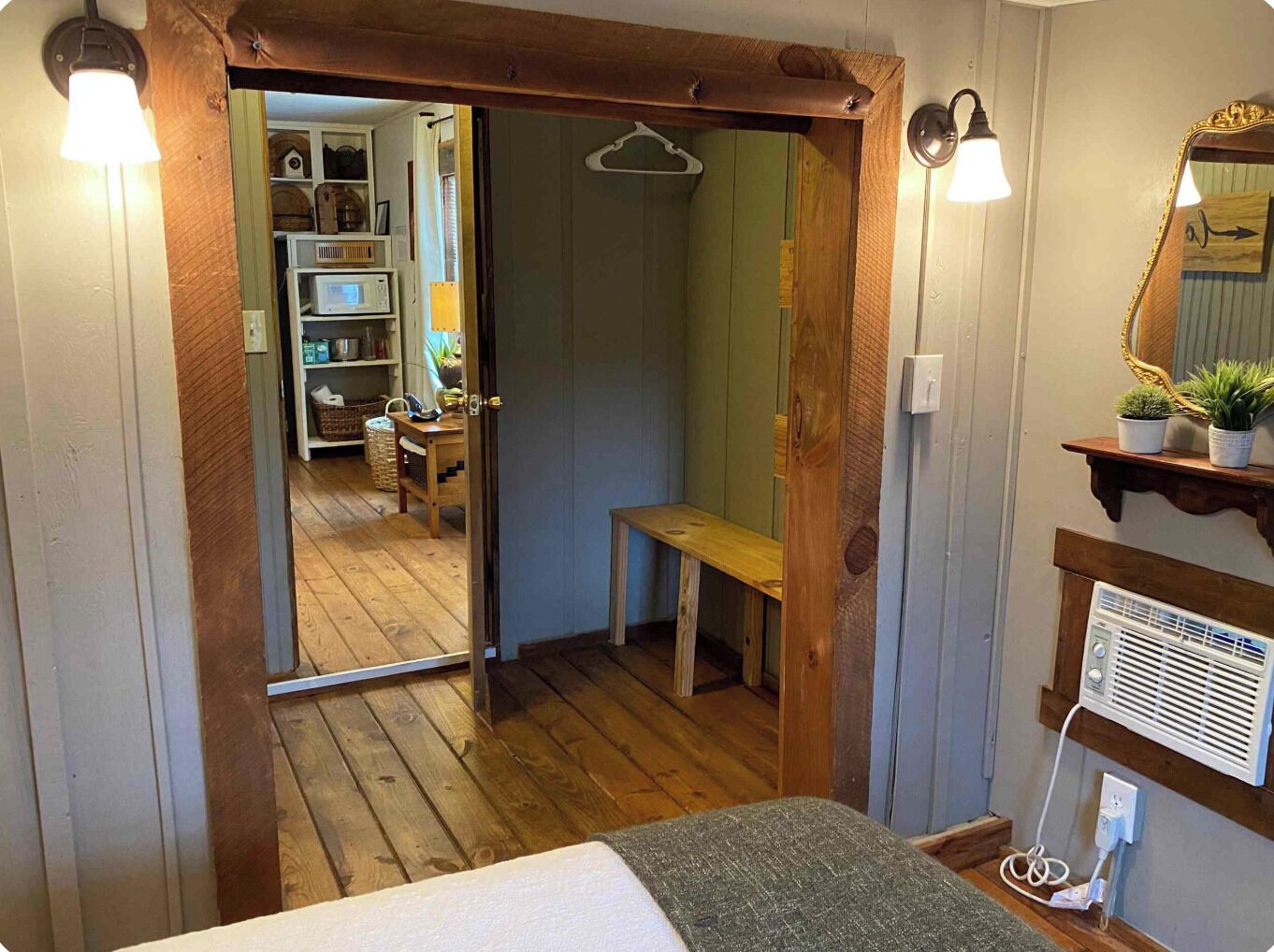 A room with a wooden door leading to a bathroom, a bench, a mirror with a decorative frame, and a shelf with potted plants.