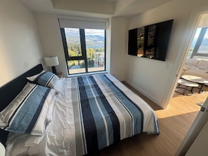 A bedroom with a large bed, a window with a view of the outdoors, a mounted TV, and a small table with chairs. - 909 Aqua 1 Resort Condo | Panoramic Okanagan Views (Kelowna)