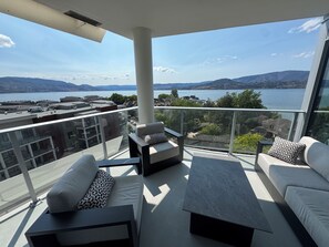 A balcony with a sofa and a table, overlooking a lake and mountains. - 909 Aqua 1 Resort Condo | Panoramic Okanagan Views (Kelowna)