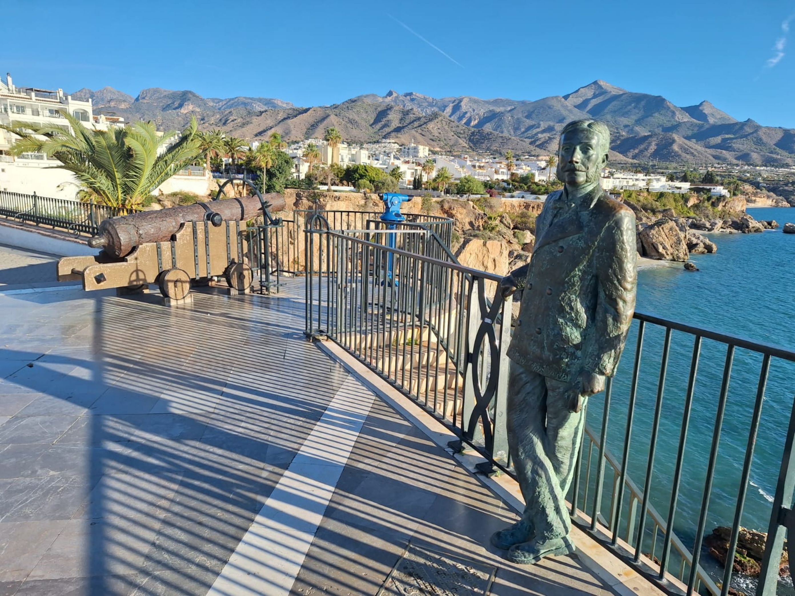 Eine Küstenpromenade mit einer Statue, einer Kanone und Blick auf Berge und eine Stadt.
