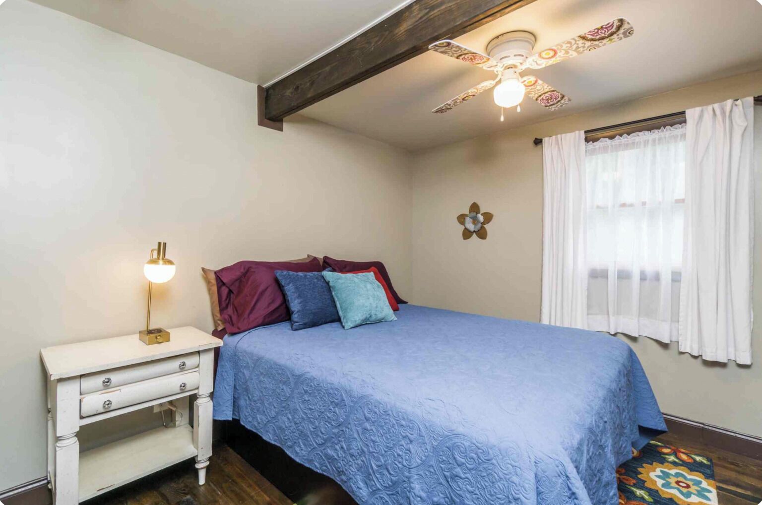Une chambre à coucher avec un lit, une table de chevet avec une lampe, un ventilateur de plafond et une fenêtre avec des rideaux.