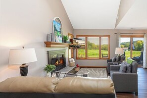 A living room with a fireplace, a sofa, a coffee table, and a view of a garden through large windows. - Perfect Fall Getaway: charming 1-bedroom townhome in Galena on golf course (Galena)