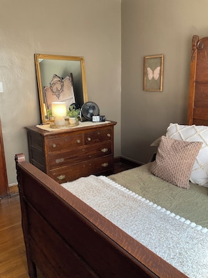 A bedroom with a wooden bed, a dresser with drawers, a mirror, a fan, and a framed butterfly picture on the wall.