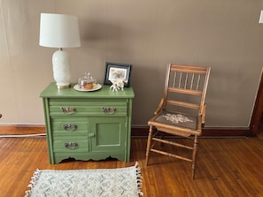 A green wooden dresser with a white lamp, a glass container, and a framed picture. Next to it is a wooden chair with a cushion.