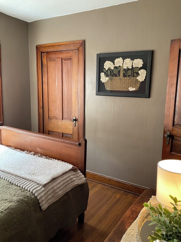 Un dormitorio con puerta de madera, una cama con manta blanca, una obra de arte enmarcada de flores y una lámpara de noche.