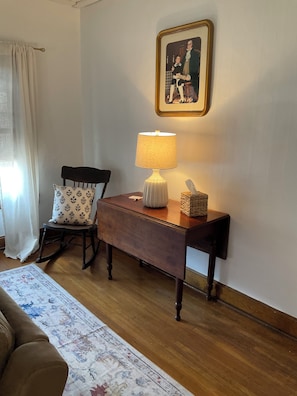 A room with a wooden desk, a rocking chair, a lamp, a framed picture on the wall, and a patterned rug.