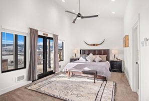 Un dormitorio con cama, ventilador de techo, ventana grande con cortinas y vistas al exterior.