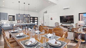 Un vaste espace de vie comprenant un coin repas avec une table pour six personnes, une cuisine équipée d’appareils électroménagers modernes et un grand téléviseur à écran plat fixé au mur.
