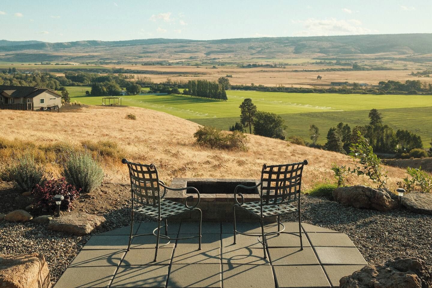 Une terrasse avec deux chaises et une table, d’où l’on peut admirer un paysage rural avec des champs et une maison au loin.