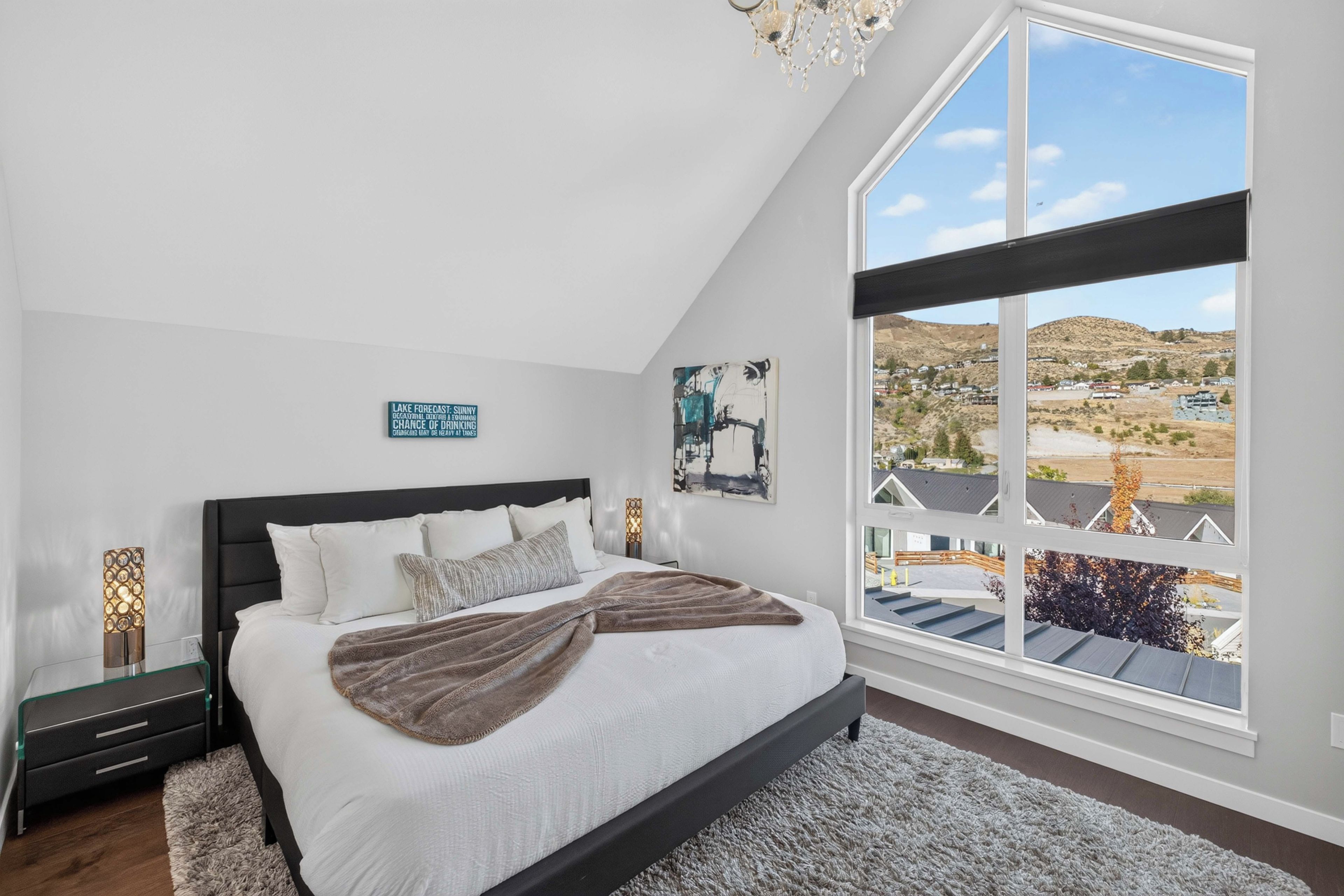 A bedroom with a large window, a bed with white bedding, a dark headboard, and a chandelier.