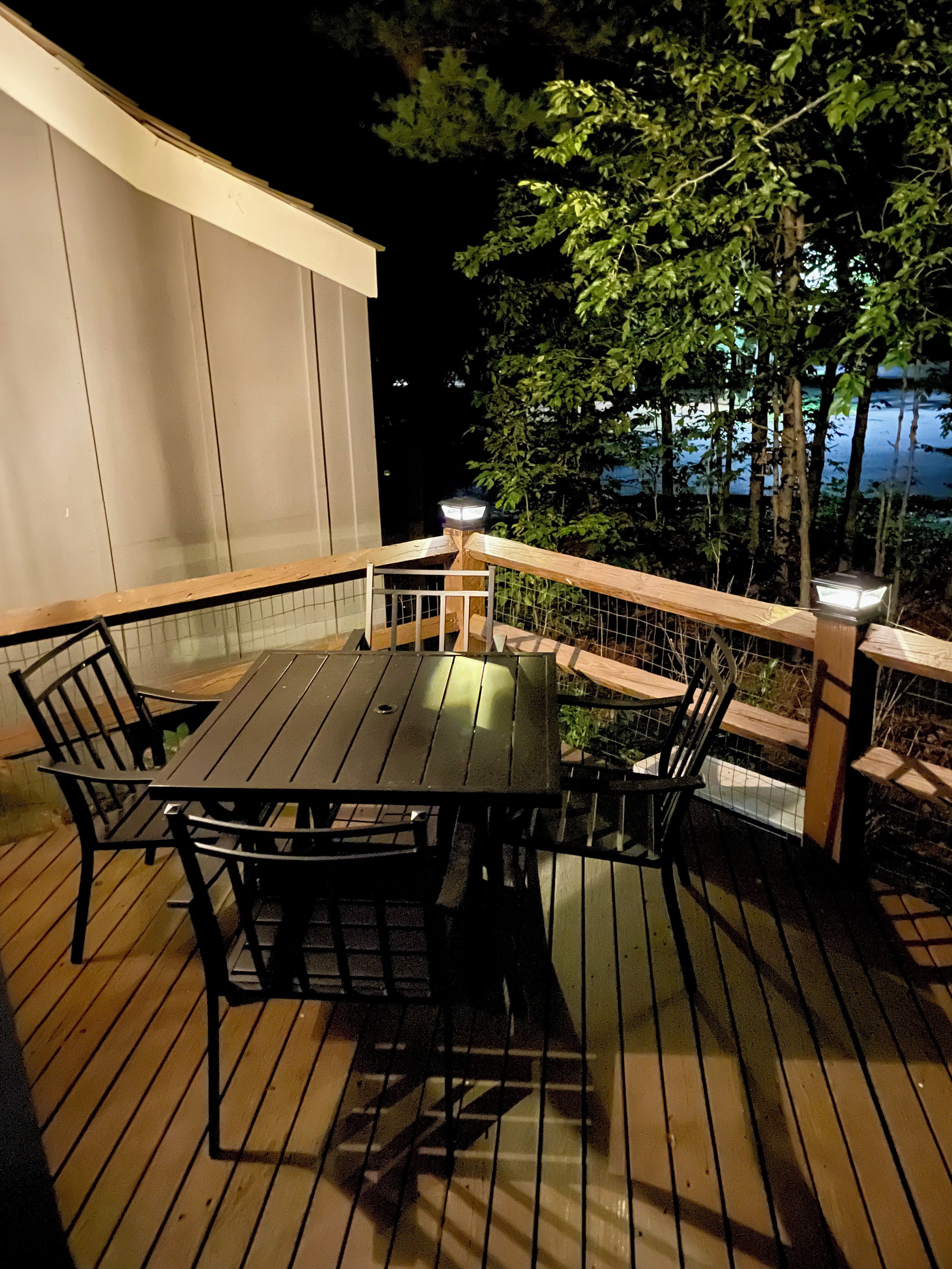 Eine Terrasse im Freien mit Tisch und Stühlen, umgeben von einem Holgitter und Blick auf Bäume sowie ein Gewässer.